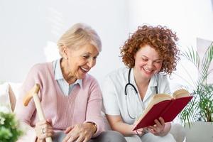 donna badante lettura un' libro mentre seduta con contento anziano donna a assistenza infermieristica casa. contento Sambuco donna seduta su bianca divano e ascoltando per infermiera lettura un' libro su forte foto