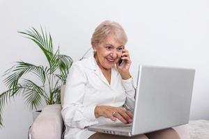 anziano donna Lavorando su il computer portatile computer, sorridente, parlando su il Telefono. anziano donna utilizzando il computer portatile. anziano donna seduta a casa, utilizzando il computer portatile computer e parlando su sua mobile Telefono, sorridente. foto