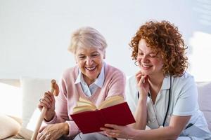 donna badante lettura un' libro mentre seduta con contento anziano donna a assistenza infermieristica casa. contento Sambuco donna seduta su bianca divano e ascoltando per infermiera lettura un' libro su forte foto