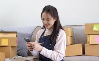 felice giovane donna asiatica imprenditrice, sorriso per il successo delle vendite dopo aver controllato l'ordine dal negozio online in uno smartphone a casa, concetto di attività commerciale online ed e-commerce foto