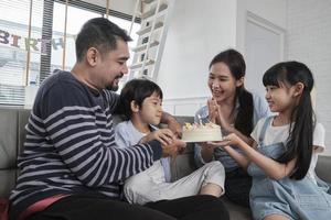 contento asiatico tailandese famiglia, giovane figlio è sorpreso con compleanno torta, regalo, colpi su candela, e celebra festa con genitori insieme nel vivente camera, benessere domestico casa evento stile di vita. foto