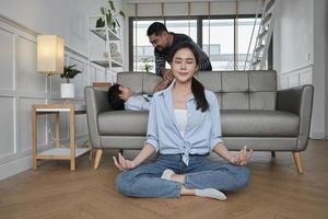 giovane asiatico tailandese madre si siede su vivente camera pavimento, medita, e pratiche yoga per Salute e benessere quando marito e figlio stuzzicare insieme su divano, contento domestico casa stile di vita su famiglia fine settimana. foto