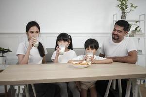 un' salutare asiatico tailandese famiglia, poco figli, e giovane genitori bevanda fresco bianca latte nel bicchiere e pane gioia insieme a un' cenare tavolo nel mattina, benessere nutrizione casa prima colazione pasto stile di vita. foto