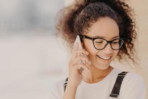 ritratto ravvicinato di donna allegra con capelli crespi, soddisfatta delle tariffe per le chiamate telefoniche, focalizzata verso il basso, tiene uno smartphone moderno, telefoni al migliore amico, discute le ultime notizie. concetto di tecnologia foto