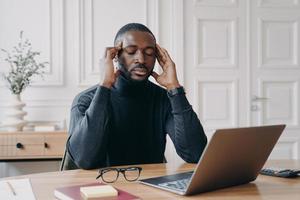 frustrato stanco uomo afroamericano impiegato con gli occhi chiusi che cerca di concentrarsi sul posto di lavoro foto