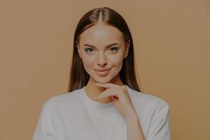 bella giovane donna europea con trucco pelle sana tocca delicatamente la mascella guarda direttamente la fotocamera ha lunghi capelli lisci bellezza naturale vestita con un maglione casual isolato su sfondo marrone foto