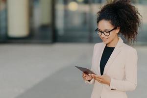 il prospero proprietario di un'azienda è in piedi con il touchpad digitale, focalizzato sullo schermo, cerca nuove idee per il mercato in via di sviluppo, indossa occhiali e giacca formale bianca. imprenditrice all'aperto foto