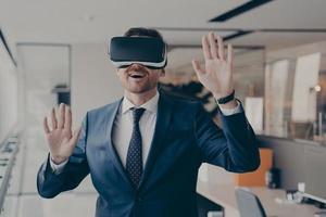 giovane uomo d'affari eccitato vestito formalmente utilizzando cuffie per realtà virtuale mentre lavorava in ufficio foto