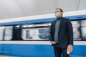 l'uomo d'affari pensieroso indossa una maschera chirurgica sterile per prevenire la diffusione di malattie respiratorie, posa contro la metropolitana, viaggia con i mezzi pubblici, tiene in mano lo smartphone. epidemia di coronavirus. foto