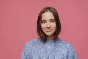 bella donna adulta con i capelli scuri, ha un'espressione del viso calma, indossa un maglione lavorato a maglia, posa in studio su sfondo rosa, pensa a qualcosa di piacevole durante l'inverno, ha un aspetto tenero foto