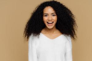 colpo alla testa di un modello femminile sano e positivo ride felice, ha folti capelli afro, vestito con un maglione morbido bianco, felice di ricevere complimenti dal fidanzato, isolato su sfondo marrone. concetto di emozioni foto