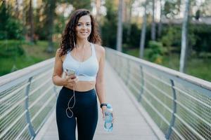 attiva bella donna sportiva in abbigliamento attivo tiene il telefono cellulare, ascolta la musica negli auricolari, si prende una pausa dopo aver fatto jogging tiene una bottiglia di acqua fresca pone al ponte all'aperto. fitness, concetto di stile di vita foto