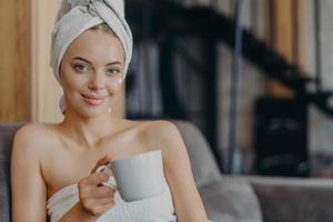 donna adorabile con trucco naturale e pelle sana, posa con le spalle nude, avvolta in un asciugamano da bagno, beve tè o caffè, si siede su un comodo divano contro l'interno della casa. persone, igiene, relax foto