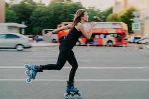 giovane in forma donna su rullo pattini con ruote Pattini durante estate giorno su occupato strada con trasporto conduce attivo stile di vita indossa nero vestiti sportivi respira fresco aria. movimento concetto. foto