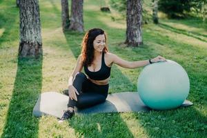 lo scatto all'aperto di una donna sportiva attiva si prende una pausa dopo gli esercizi fisici, posa sul tappetino fitness con fitball, gode dell'aria fresca nella foresta, ha una forma corporea perfetta utilizza attrezzature sportive. concetto di ginnastica foto