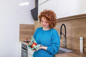 carino ragazza con Riccio rosso capelli sferzata sua Mela con cucina asciugamano. giovane donna pulizia sua frutta nel sua cucina. quotidiano presa di vitamine con frutta, dieta e salutare mangiare foto