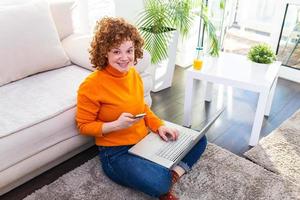 carino femmina rosso capelli libero professionista lettura testo messaggi mentre seduta su pavimento con Aperto computer nel moderno casa, giovane creativo donna opera su il computer portatile mentre avendo arancia succo foto