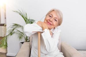 sorridente nonna seduta su divano. ritratto di un' bellissimo sorridente anziano donna con a piedi canna su leggero sfondo a casa. vecchio donna seduta con sua mani su un' canna foto