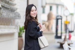 ritratto di una bella donna asiatica dai capelli lunghi con un cappotto nero, che porta una borsa da passeggio, sorridente di buon umore in città all'aperto. foto