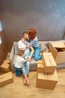 giovane contento famiglia con ragazzo disimballaggio scatole insieme seduta su divano foto