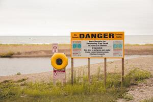 Pericolo cartello su sabbioso spiaggia di lago Michigan. erbacce in crescita nel il sabbia. nuvoloso nuvoloso cielo. ciottoli misto nel il sabbia. avvertimento segni postato su il spiaggia. onde nel il lontano acqua. foto