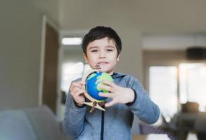 ritratto ragazzo è guardare a un' globo, scuola ragazzo apprendimento di geografia, giovane ragazzo con contento viso Tenere mondo carta geografica in piedi nel vivente camera, mondo bambini S giorno, formazione scolastica e homeschooling concetto foto