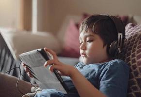 autentico ritratto ragazzo seduta su divano Guardando cartoni animati su tavoletta, yong ragazzo giocando gioco su toccare pad, bambino dire bugie su divano indossare cuffie ascoltando per musica o rilassante su il suo proprio nel vivente camera foto