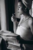 bellissimo giovane donna nel un' asciugamano fuma un' sigaretta e legge giornale foto