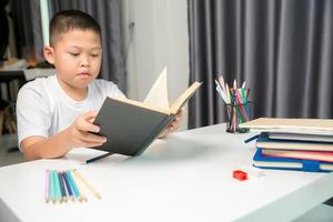 ragazzo persona seduta studia interno a casa, maschio ragazzo alunno in linea apprendimento e fare compiti a casa su scrivania, giovane bambino lettura e scrittura un' libro su tavolo. concetto di formazione scolastica, tecnologia cyberspazio foto