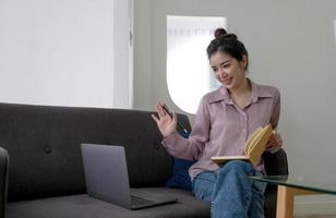 giovane allegro asiatico donna seduta su divano e Guarda per il computer portatile con video chiamata incontro e studia in linea su il Internet. foto