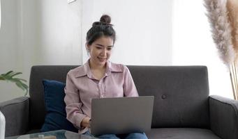 giovane donna asiatica freelance d'affari che lavora al computer portatile controllando i social media mentre è sdraiata sul divano quando si rilassa nel soggiorno di casa. stile di vita latino e donne ispaniche di etnia al concetto di casa. foto