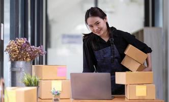 giovane attraente donna asiatica proprietario avvio affari guardare fotocamera lavoro felice con scatola a casa preparare la consegna pacchi nella catena di approvvigionamento delle PMI foto