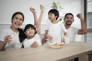 un' salutare asiatico tailandese famiglia, poco figli, e giovane genitori bevanda fresco bianca latte nel bicchiere e pane gioia insieme a un' cenare tavolo nel mattina, benessere nutrizione casa prima colazione pasto stile di vita. foto