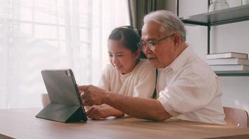 un asiatico nonno e nipotina la spesa tempo insieme nel il vivente camera. contento anziano uomo con poco ragazza utilizzando un' tavoletta su il tavolo a casa. foto