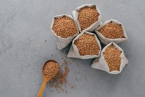 vista dall'alto del grano saraceno marrone in sacchi e cucchiaio di legno su sfondo grigio. concetto di dieta sana. semole di grano saraceno. pianta biologica coltivata biologicamente foto