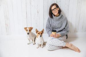 tiro al coperto di una bella donna che indossa occhiali e abbigliamento casual, beve bevande calde, si gode il buongiorno, si siede vicino ai suoi animali domestici preferiti su uno sfondo di recinzione bianca. concetto di felicità e tempo libero foto