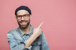 colpo orizzontale di allegro uomo barbuto con espressione felice, vestito con abiti alla moda, indossa occhiali, cappello e camicia di jeans, indicato con il dito anteriore nello spazio vuoto della copia per la tua pubblicità foto