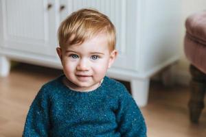 vicino su ritratto di bionda poco bambino con paffuto guance, sembra con blu occhi in telecamera, ha innocente Guarda. adorabile infantile ha grave espressione si siede al di sopra di casa interno. infanzia concetto foto