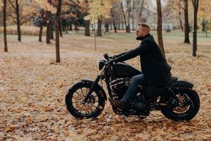 inquadratura laterale di un motociclista maschio sicuro di sé che guida la moto, ha un'avventura su due ruote nel parco autunnale, indossa abiti eleganti, occhiali da sole protettivi, si gode la natura durante la bella stagione autunnale. foto