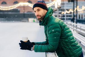 un uomo premuroso con una folta barba si sporge da qui, tiene il caffè da asporto, guarda la partita di hockey durante l'inverno, pensa a qualcosa. il maschio sognante con la barba lunga beve una bevanda calda quando fa freddo foto