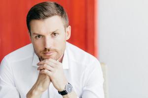 inquadratura di un uomo attraente e premuroso con la barba lunga tiene il mento, vestito con un'elegante camicia bianca, aspetta qualcuno, essendo un uomo d'affari prospero, posa su sfondo bianco e rosso con spazio libero da parte foto