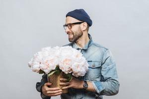 il colpo orizzontale del maschio barbuto felice distoglie lo sguardo mentre tiene un mazzo di fiori bianchi, essendo di buon umore come ha appuntamento con la sua ragazza, le fa un regalo, isolato su sfondo bianco. foto