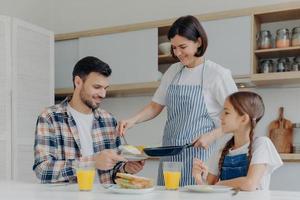 la casalinga indaffarata tiene la padella, dà un pasto preparato a marito e figlia, fa una deliziosa colazione insieme, si siede al tavolo della cucina con un bicchiere di succo e un panino. la famiglia felice mangia a casa foto