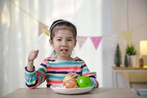 bambino asiatico ragazza mangiare Mela su il scrivania, salutare cibo abitudini concetto foto