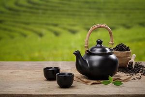 tazza di caldo tè con teiera, verde tè le foglie e secco erbe aromatiche su il di legno tavolo nel piantagioni sfondo con vuoto spazio, biologico Prodotto a partire dal il natura per salutare con tradizionale foto