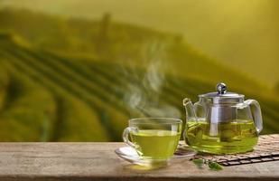 caldo tazza di verde tè e bicchiere brocche o barattoli e verde tè foglia su di legno tavolo con il tè piantagioni sfondo foto
