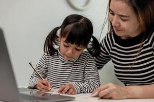 ragazzina asiatica che impara la lezione online a casa con la madre. il bambino in età prescolare usa il computer portatile per fare i compiti, scuola a casa dall'insegnante di scuola tramite Internet digitale remoto con il supporto della mamma. foto