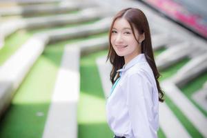 bellissimo asiatico alto scuola alunno ragazza nel il scuola uniforme con bretelle su sua denti in piedi e sorrisi con fiducia mentre lei sembra a il telecamera felicemente con il edificio nel il sfondo. foto