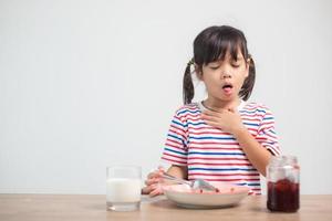 asiatico bambini ragazze mangiare pane e dolci e attaccare nel quelli gole con soffocato cibo o vomito su il tavolo a casa per prima colazione o pranzo foto