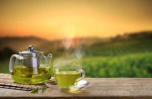 caldo tazza di verde tè e bicchiere brocche o barattoli e verde tè foglia su di legno tavolo con il tè piantagioni sfondo foto